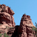 Picture of two red rock spires against an azure sky with the rising quarter moon and a jet flying up between them.